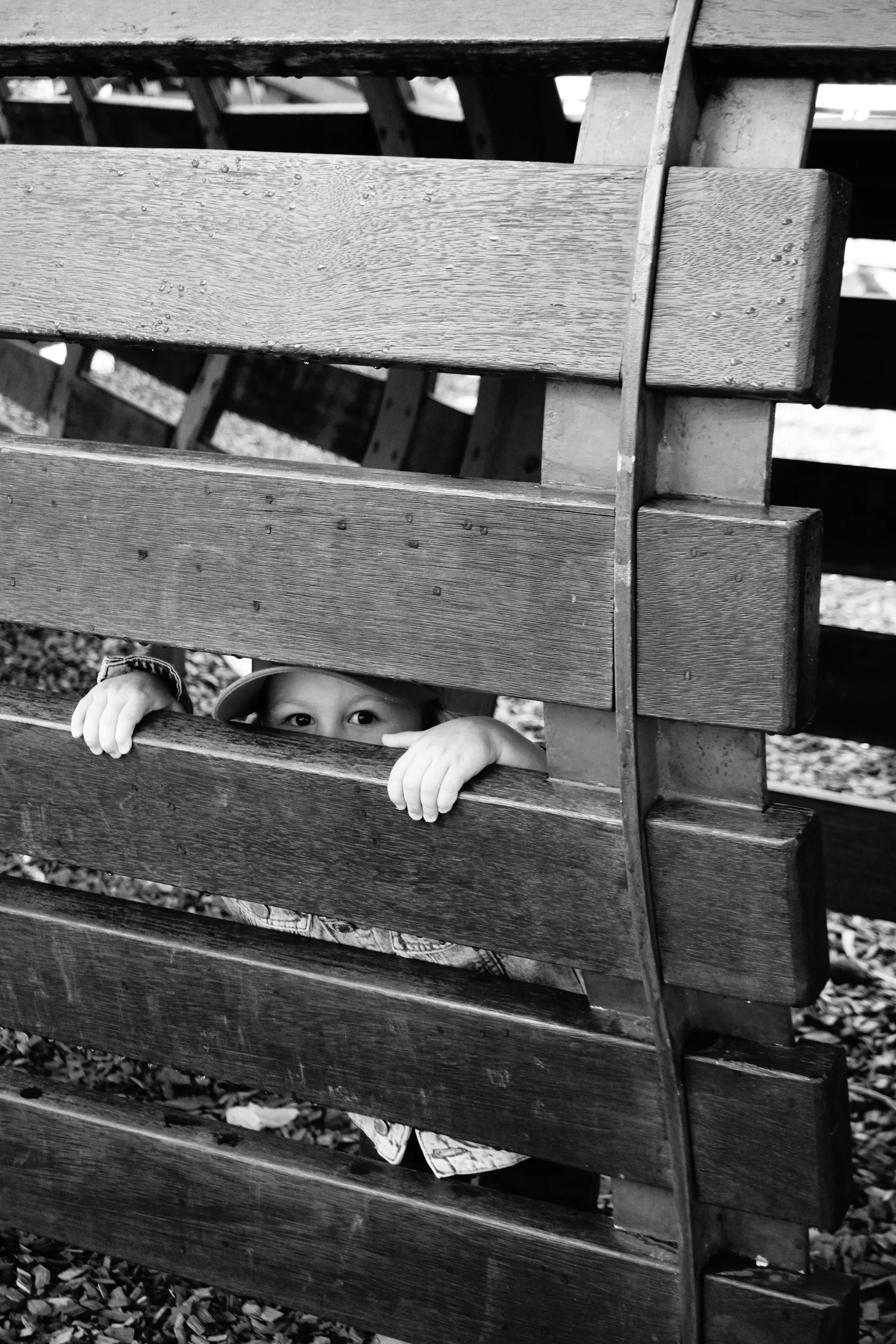 A child peeking through the horizontal wooden slats of a playground fence, with only their eyes and hands visible.
