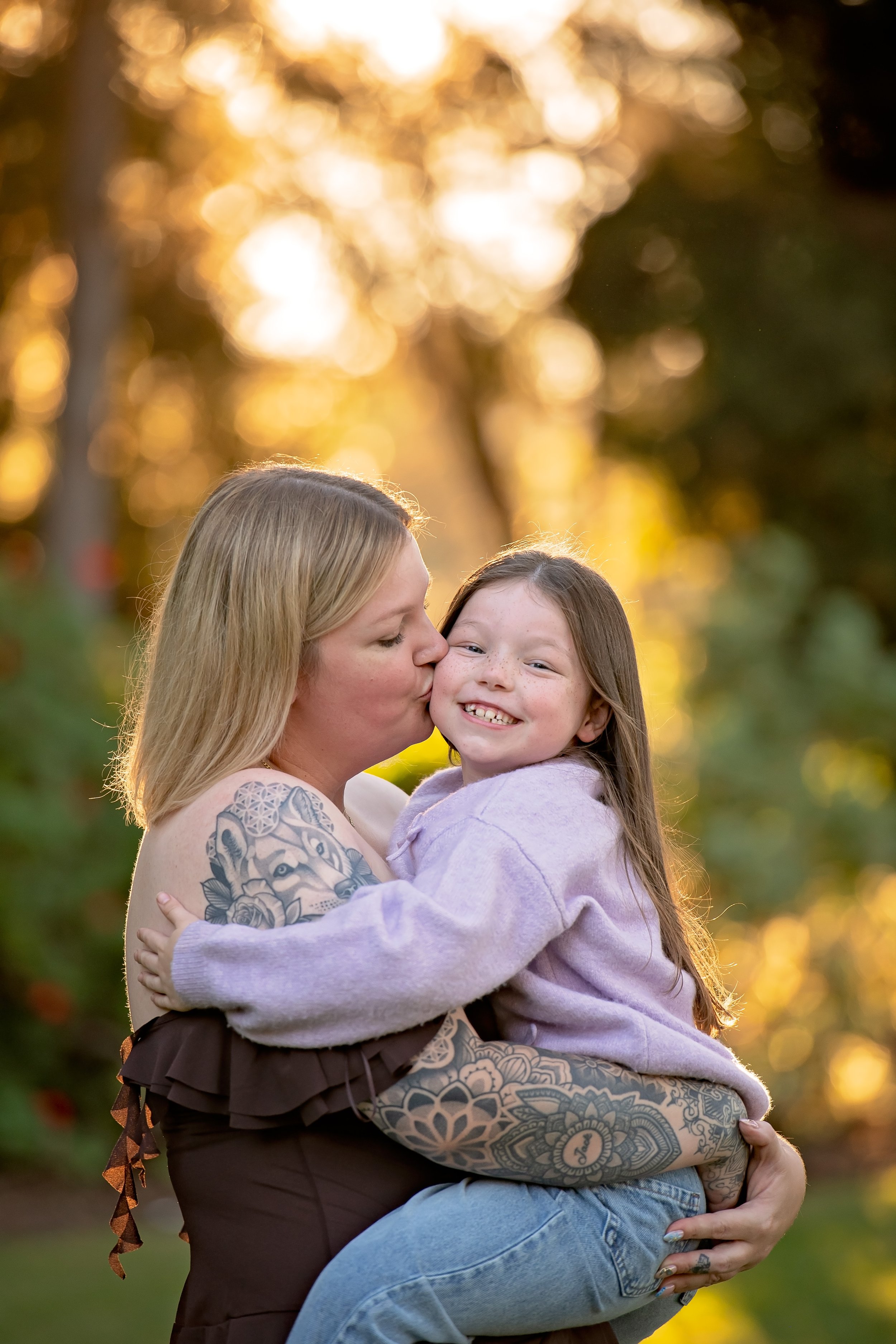 A woman with tattoos on her arm holding a smiling young girl in her arms outdoors during sunset. The woman is giving the girl a kiss on the cheek, and the background features a bokeh of trees and bright sunlight.