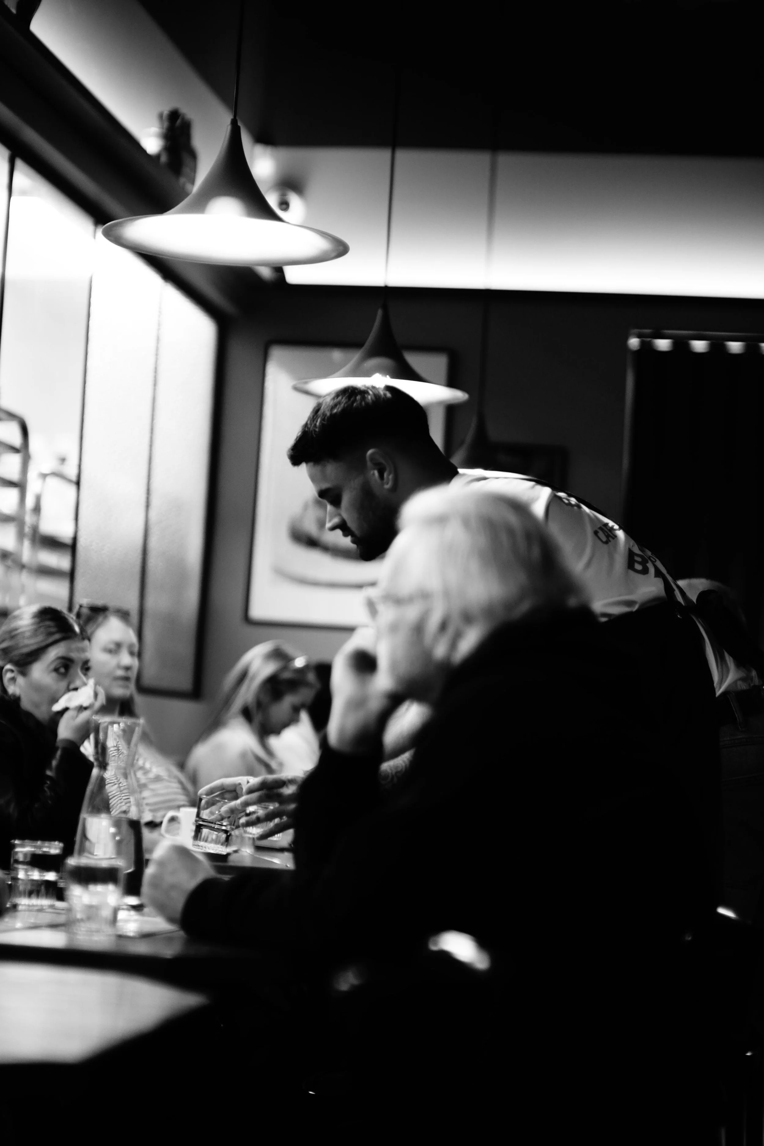 People dining in a restaurant with hanging lights, some are eating, and a waiter is serving or taking orders.