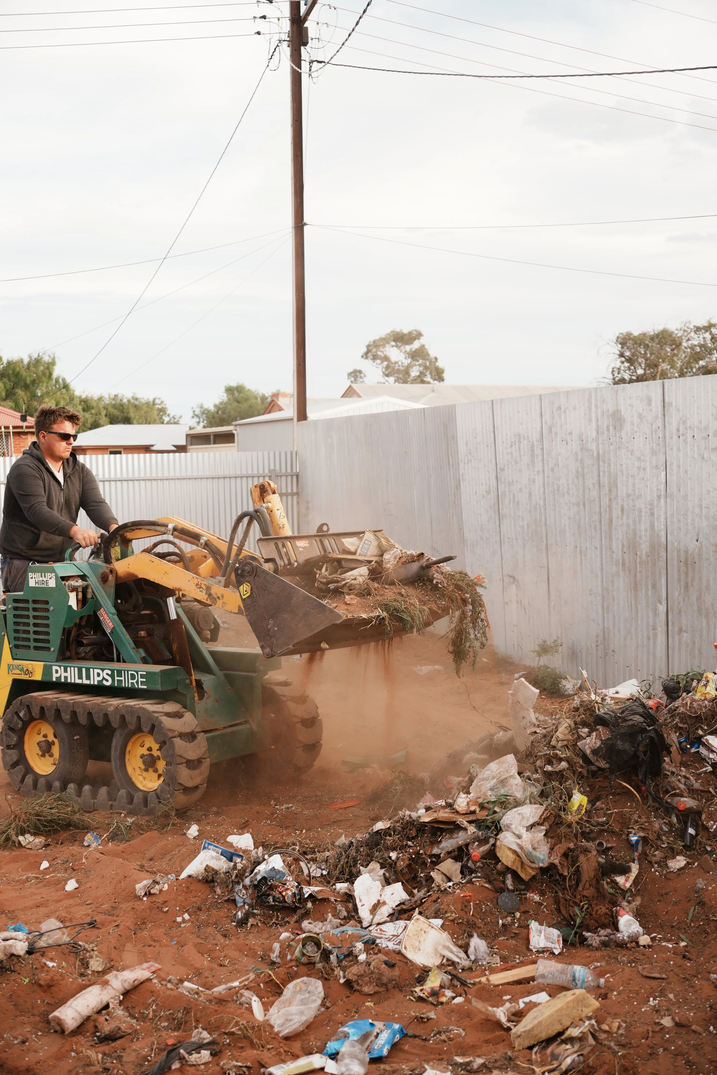 A man operating a small front loader or bulldozer clearing trash and debris in an outdoor area. Trash and debris are scattered on the ground.