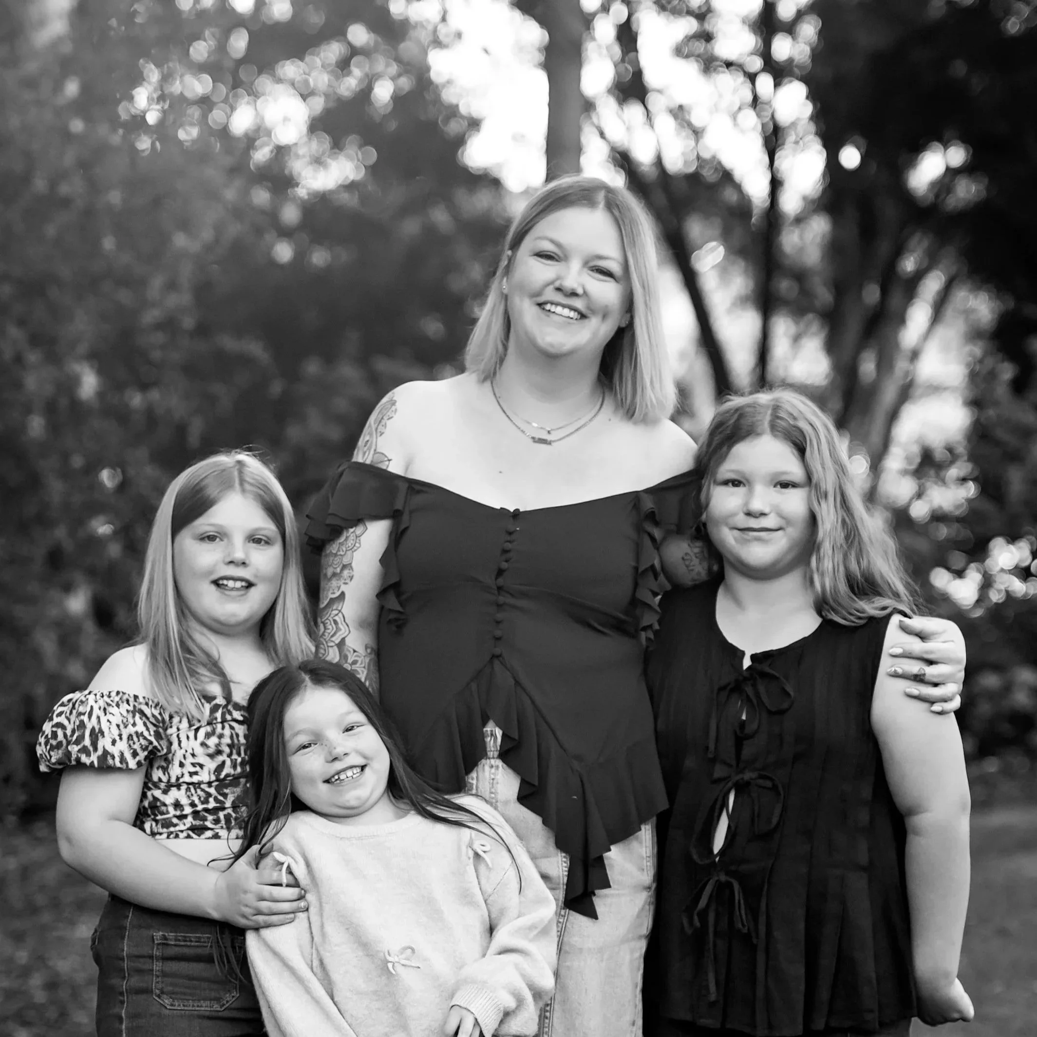 A group of young girls and one woman outdoors, smiling and posing together in a park with trees in the background, captured in black and white.