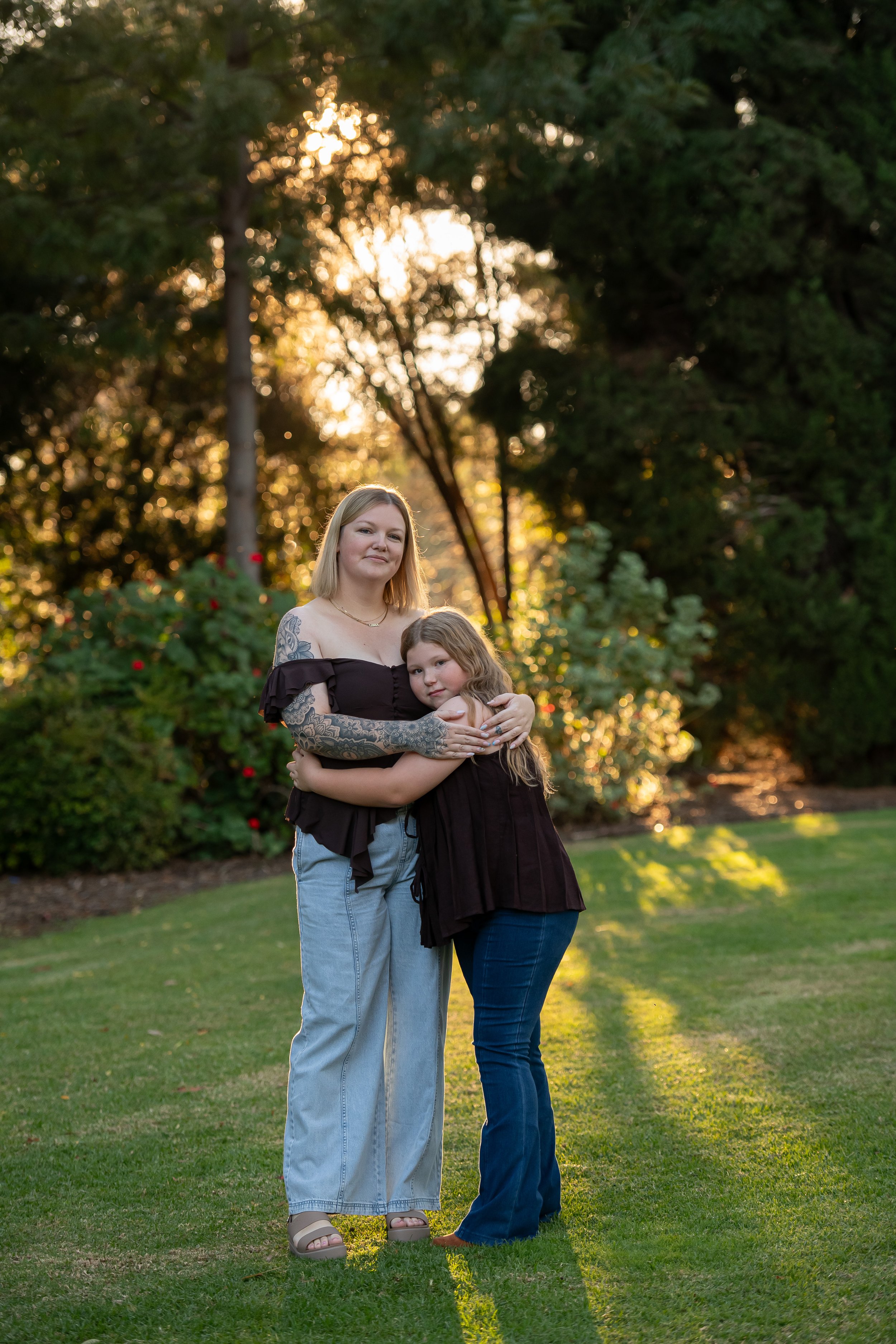 A woman with tattoos on her arm and shoulder and a young girl are standing outdoors on grass during sunset. The woman is wearing a black off-the-shoulder top and light blue jeans, while the girl is dressed in a black dress and blue jeans. They are hugging each other with trees and sunlight in the background.