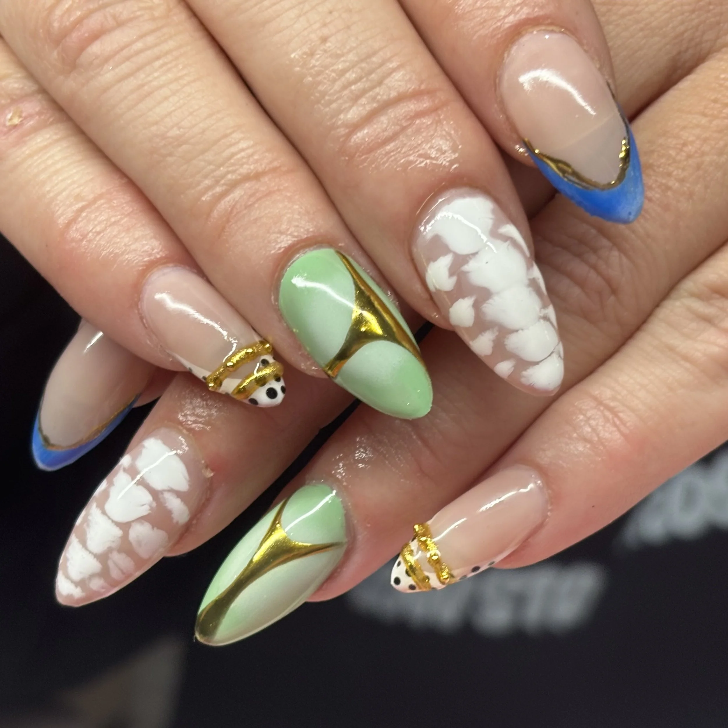 Close-up of a hand with decorated nails, featuring various colors and patterns, including white with black polka dots, pastel green, and a blue tip with gold accents.