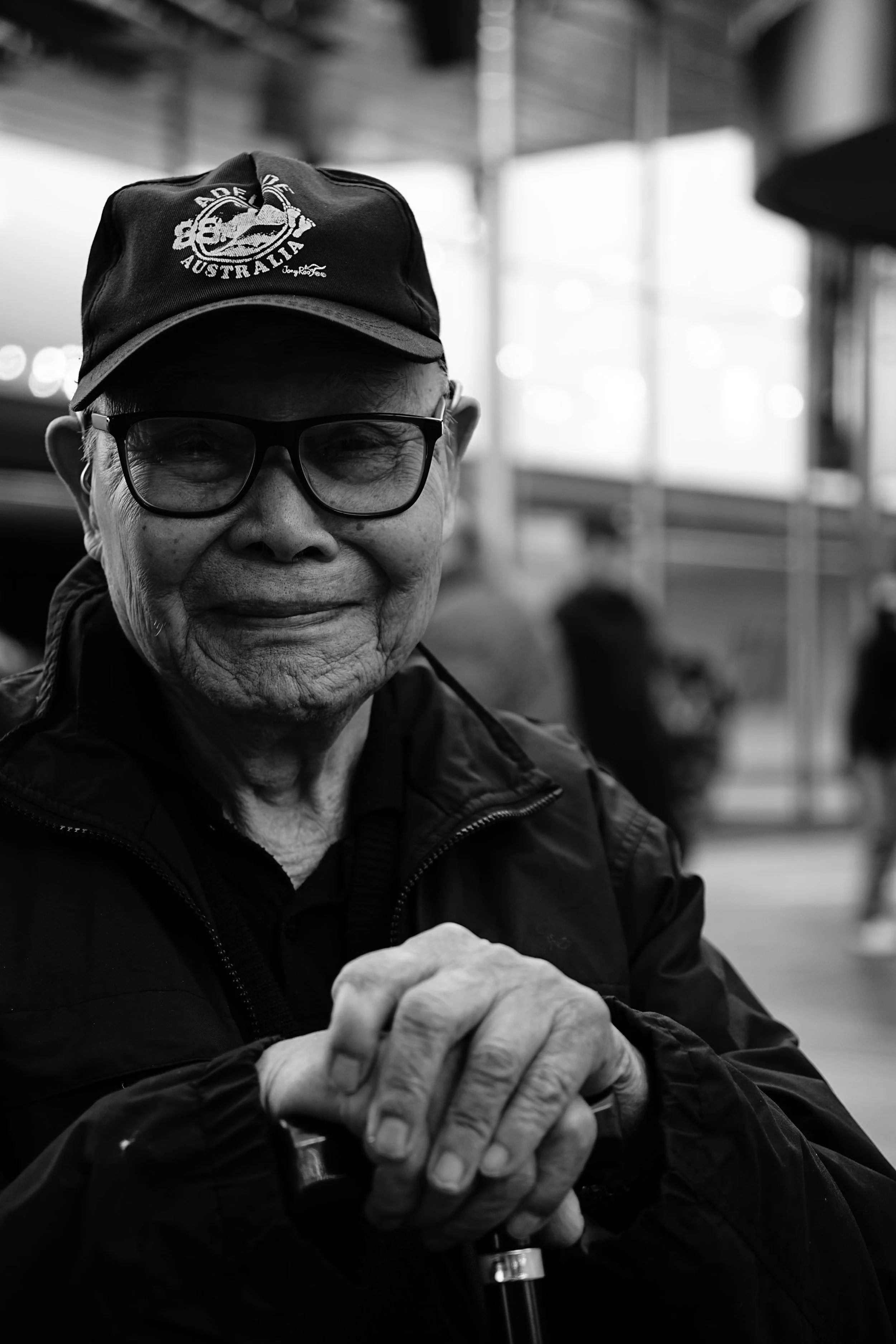 An elderly man wearing a cap with 'Aussie' written on it, glasses, and a dark jacket, smiling while clasping his hands on a cane in an indoor setting with blurred people in the background.
