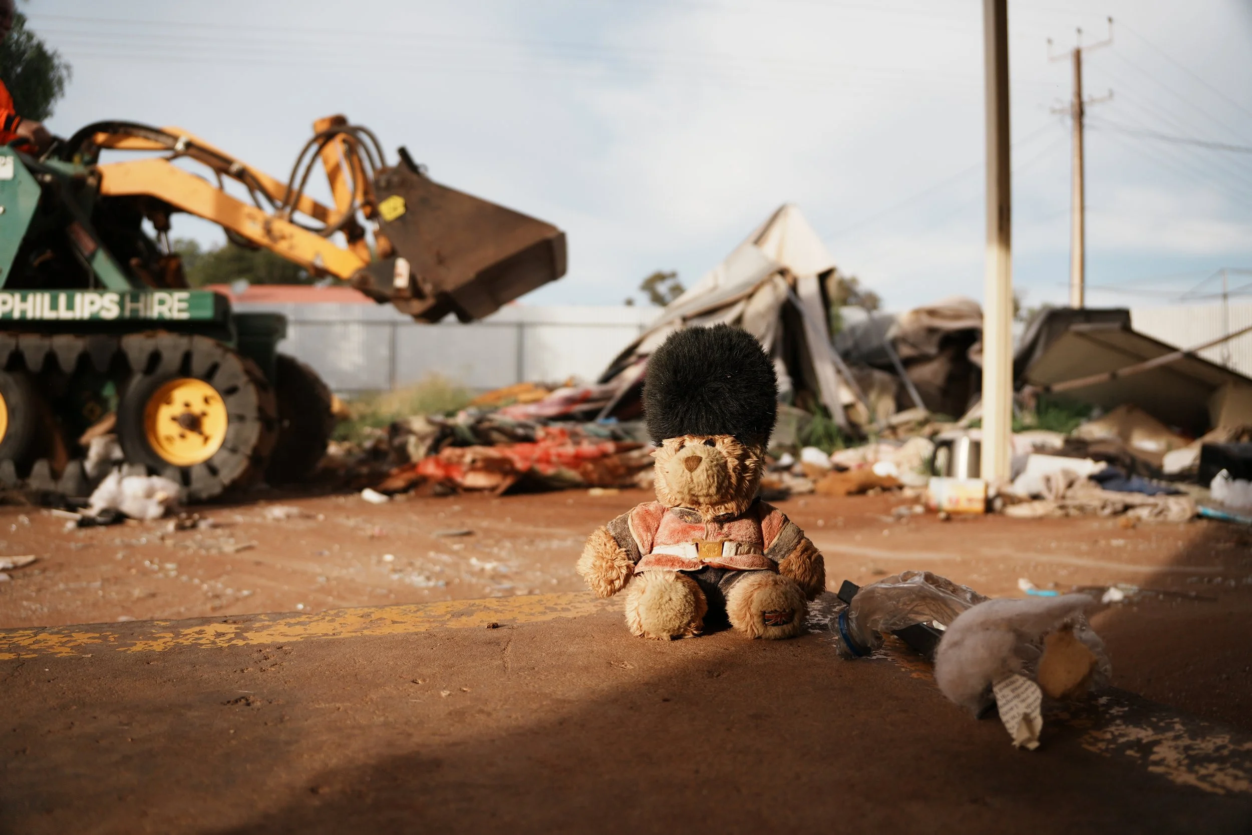 A teddy bear with a backpack sitting in front of debris and wreckage in an outdoor setting, with power lines in the background.