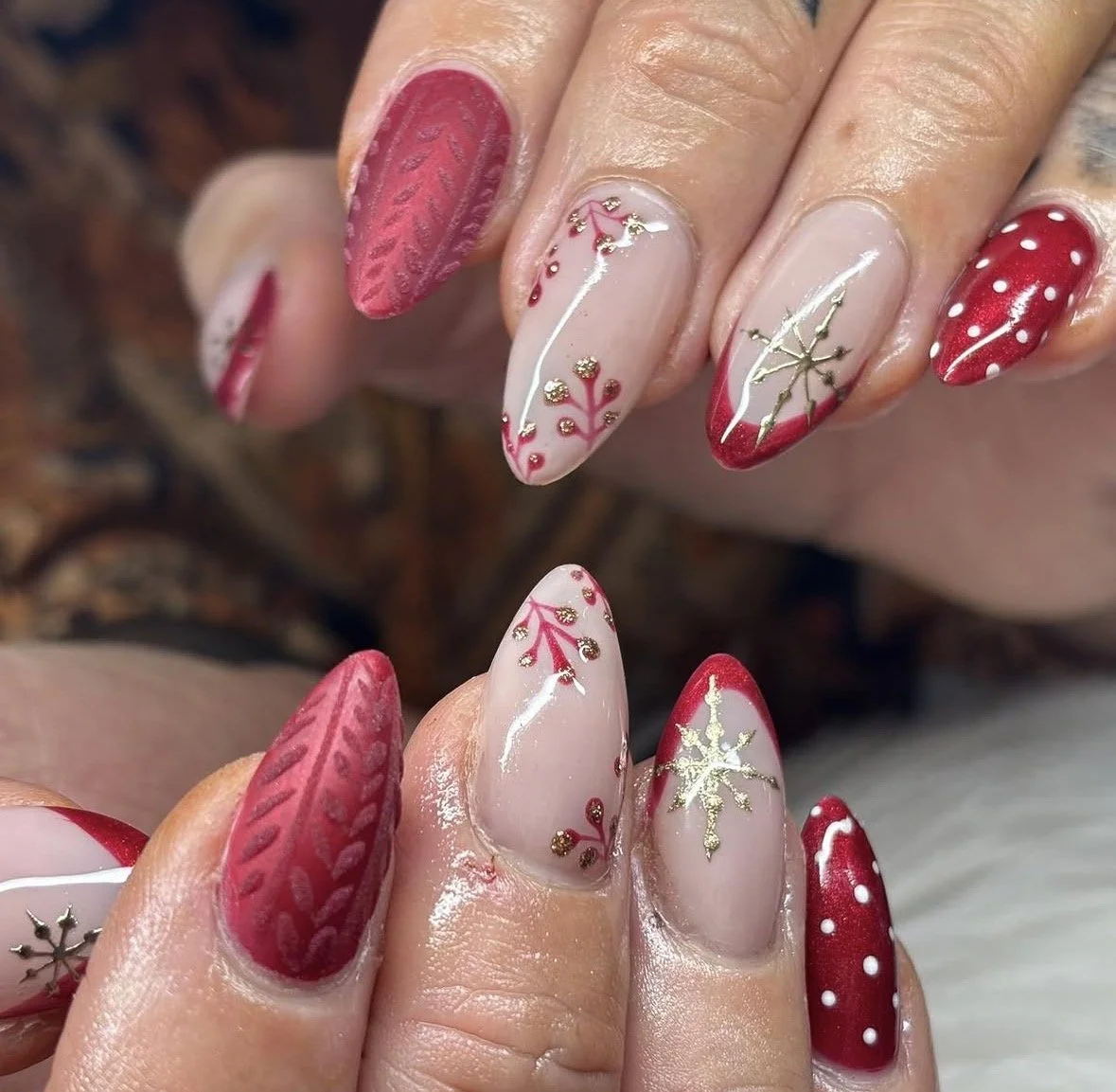 Close-up of Christmas-themed nail art with red, white, and gold designs, including snowflakes, holly, and polka dots on long almond-shaped nails.