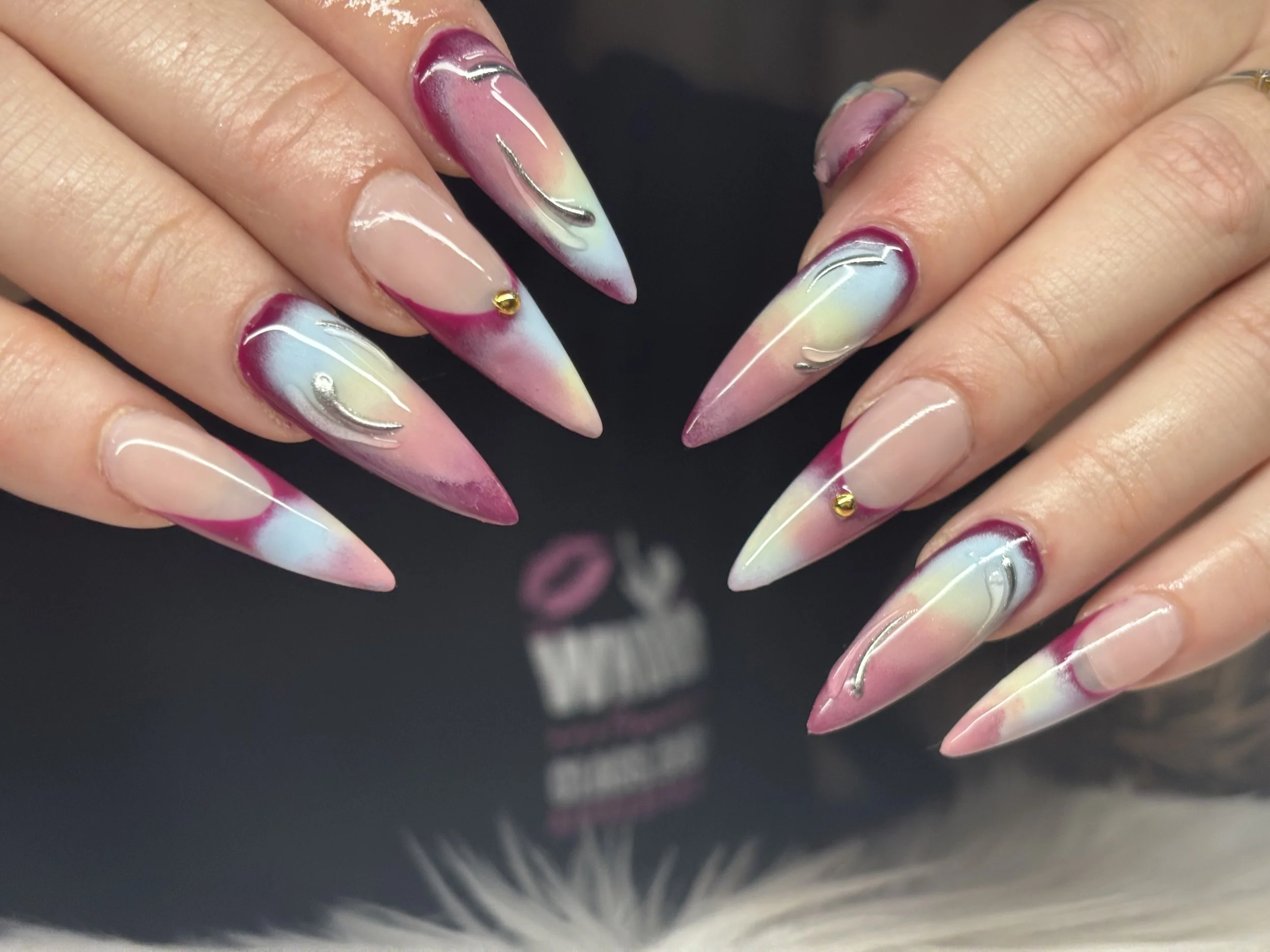Close-up of a hand with long, pointed nails painted in a pastel swirl of pink, blue, yellow, and white, decorated with thin silver lines and small gold studs.