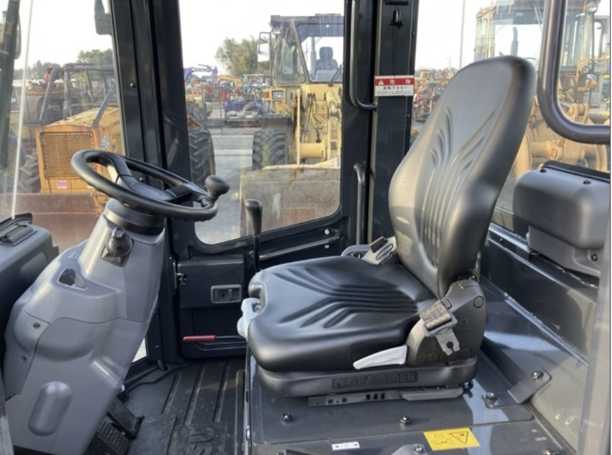 Interior view of a construction vehicle cabin with steering wheel and black seat, with construction equipment visible through the windows.