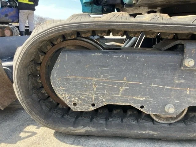 Yellow Komatsu PC30MR mini excavator parked on sidewalk next to chain-link fence with water body in background.