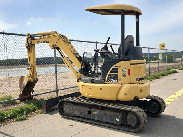 Yellow Komatsu PC30MR mini excavator parked on sidewalk next to chain-link fence with water body in background.