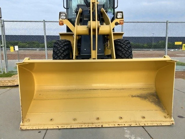 Front view of a yellow construction loader with a large bucket in the foreground, parked on pavement with a chain-link fence in the background.