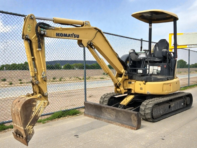 A Komatsu mini excavator parked on a paved surface near a chain-link fence with a body of water and green landscape in the background.