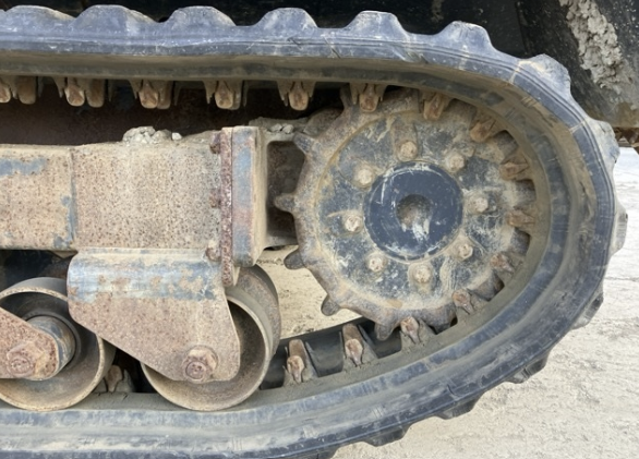Close-up of a tracked construction vehicle's undercarriage, showing a worn rubber track and metal components with dirt and rust.