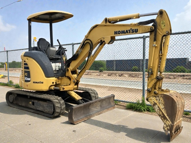 A yellow Komatsu mini excavator with a digging bucket, parked on a paved surface next to a chain-link fence, under a partly cloudy sky.