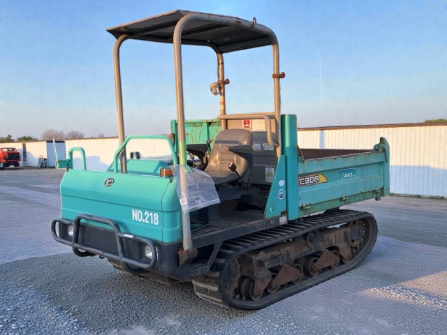A small blue and black tracked construction vehicle with an open driver seat and a canopy, parked on gravel with a white fence and a few trucks in the background.