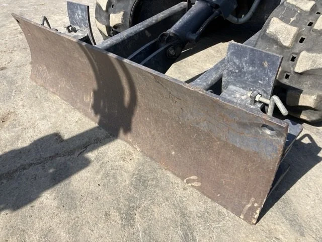Small yellow Komatsu mini excavator with a protective canopy, parked on a concrete sidewalk next to a chain-link fence, with a dirt area and cloudy sky visible in the background.