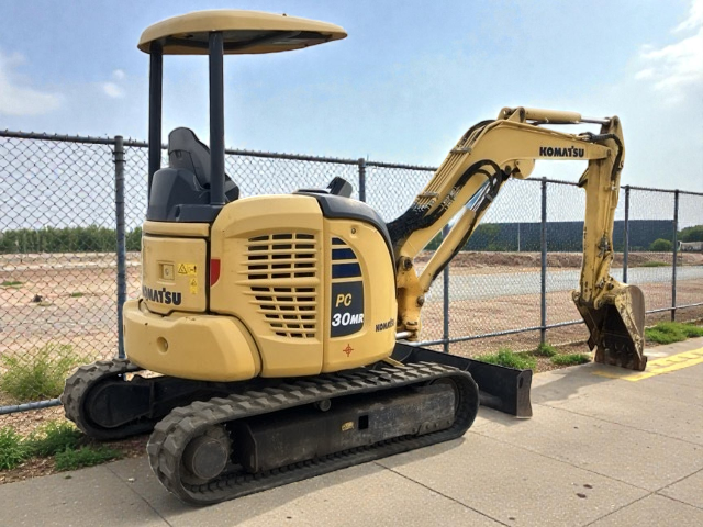 Small yellow Komatsu PC30MR mini excavator with a black bucket, parked on a concrete surface next to a chain-link fence with a dirt area and blue sky in the background.
