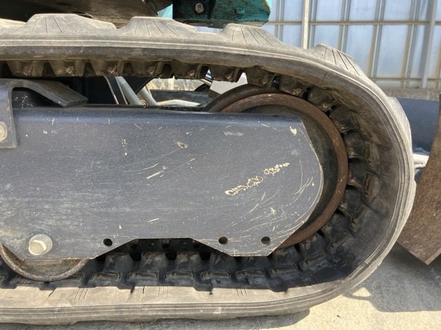 Close-up view of a large excavator bucket attached to an arm, positioned on concrete ground outdoors, with a chain-link fence, pavement, and some trees and buildings in the background.