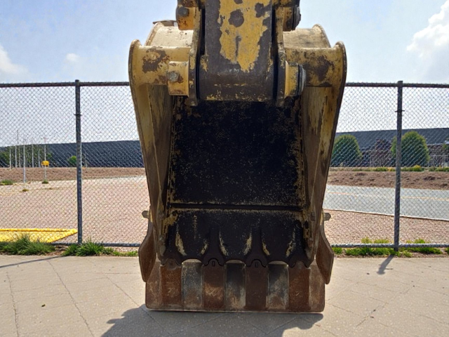 Close-up view of a large excavator bucket attached to an arm, positioned on concrete ground outdoors, with a chain-link fence, pavement, and some trees and buildings in the background.