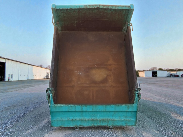 Open turquoise dump trailer with empty bed in an industrial area with multiple warehouses.