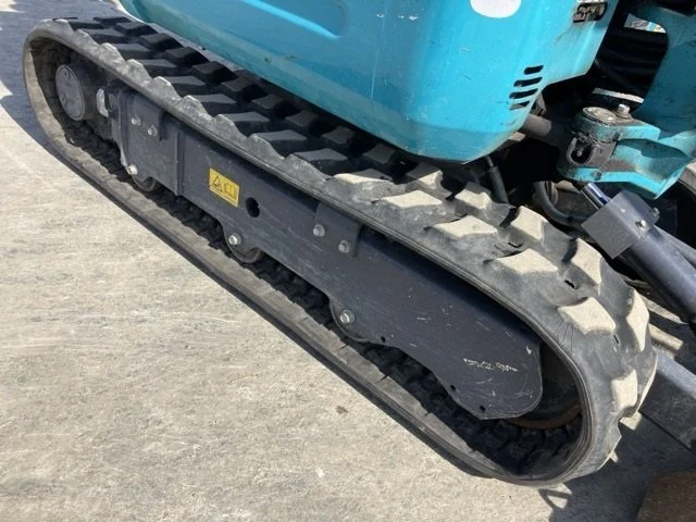 A yellow Komatsu mini excavator with a digging bucket, parked on a paved surface next to a chain-link fence, under a partly cloudy sky.