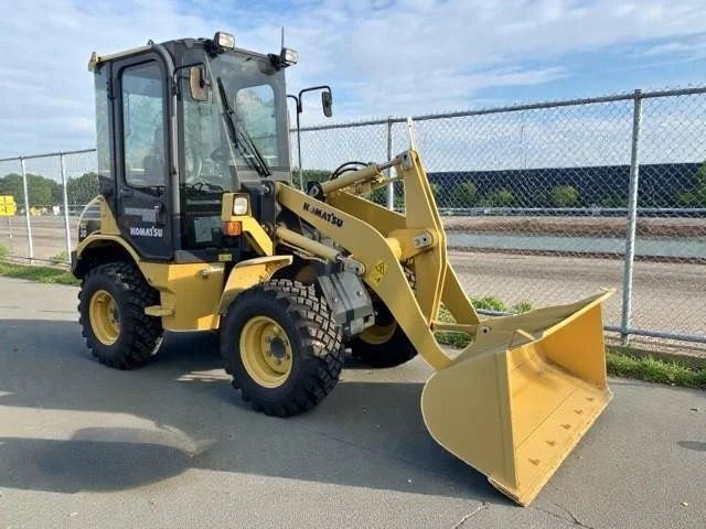 Yellow Komatsu front loader with a bucket attachment parked on a paved surface next to a chain-link fence, with a partly cloudy sky in the background.
