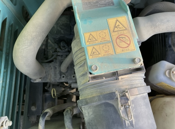 A close-up view of a dusty industrial machinery component with warning labels, hoses, and metal parts.