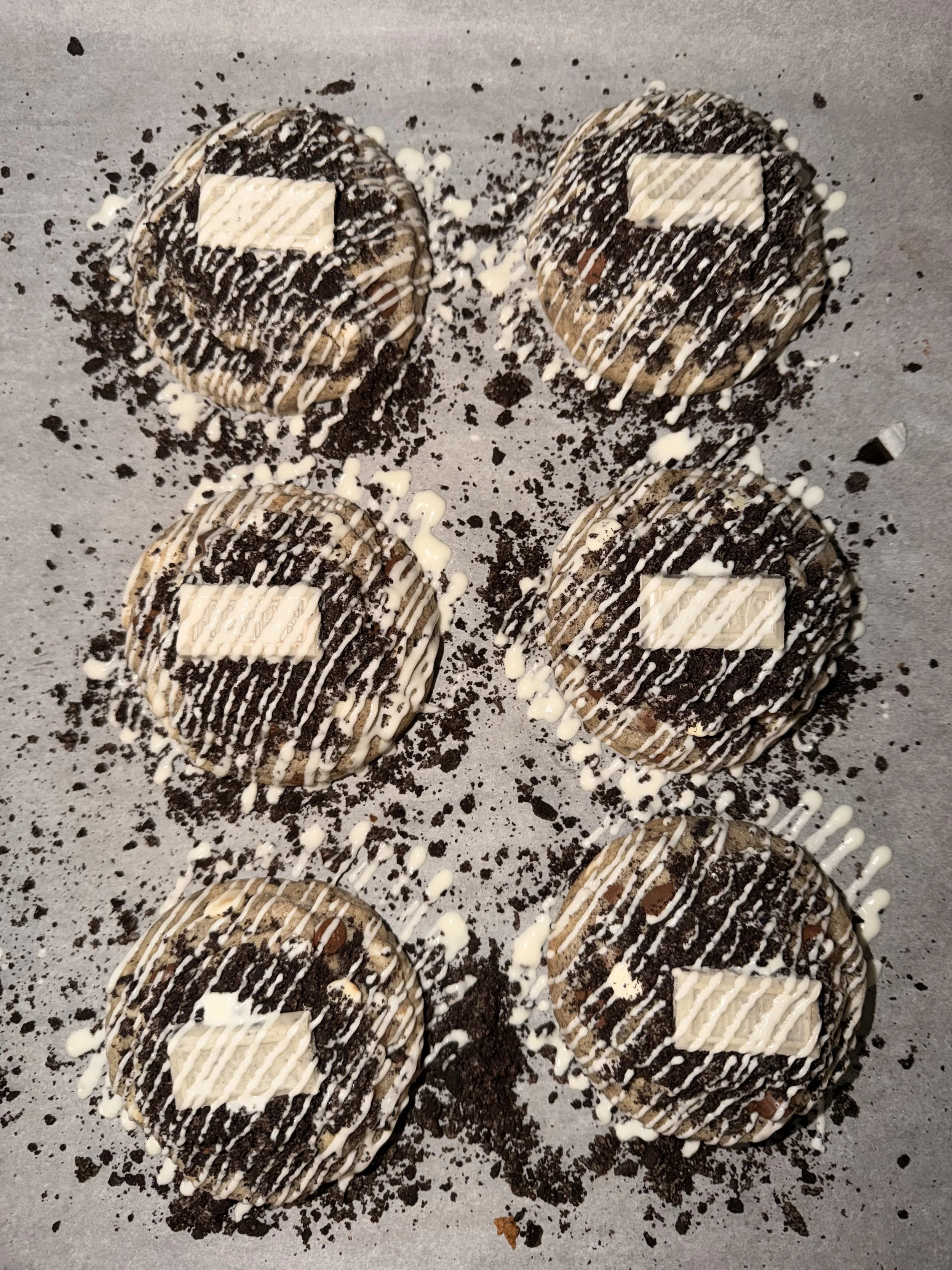 Six cookies decorated with white and dark chocolate drizzle, crumbled Oreos, and white chocolate squares, arranged on parchment paper with cookie crumbs scattered around.