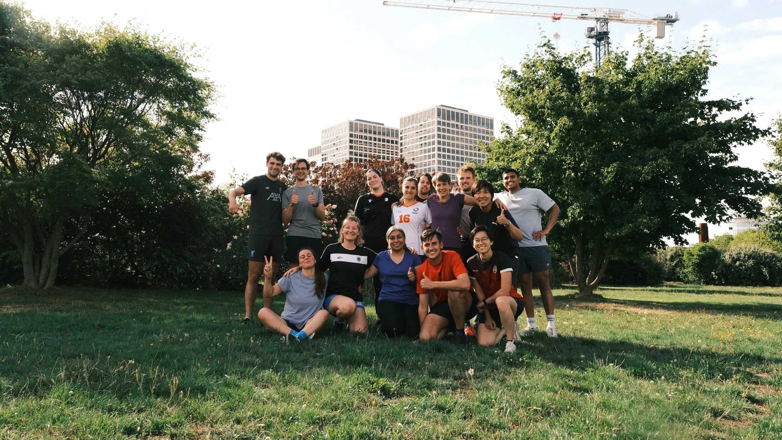 Group of people smiling and posing together outdoors on a grassy area with trees and buildings in the background, some making peace signs or thumbs up.