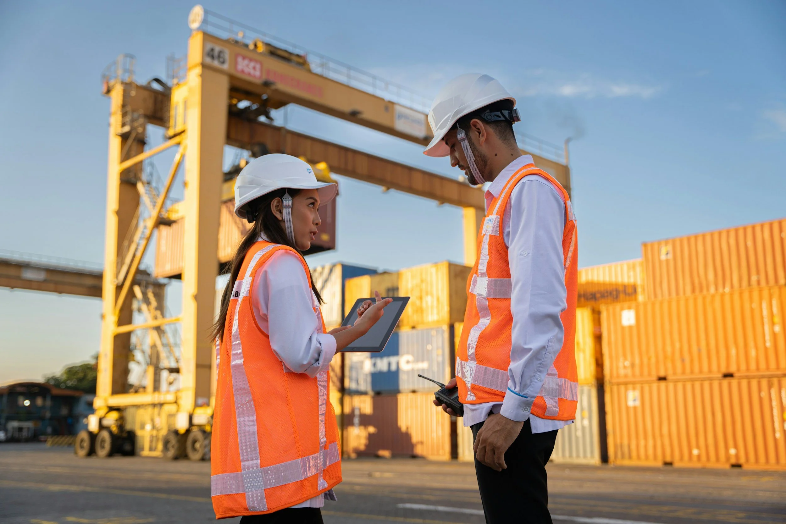 Two workers in safety helmets and orange safety vests standing in front of shipping containers and a crane at a port, discussing using a tablet and radio devices during the daytime.