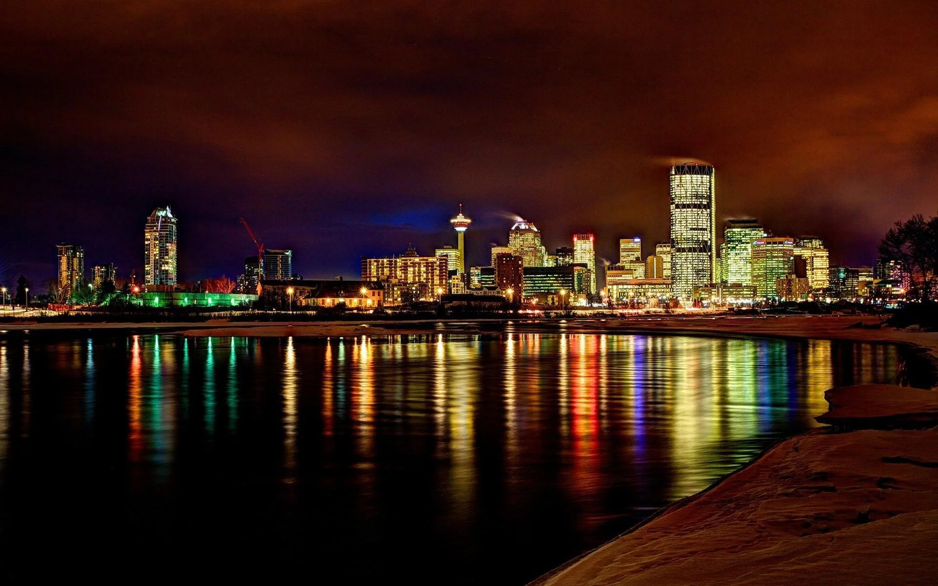 Night view of a city skyline with illuminated skyscrapers and reflections on the water, with snow-covered ground in the foreground.