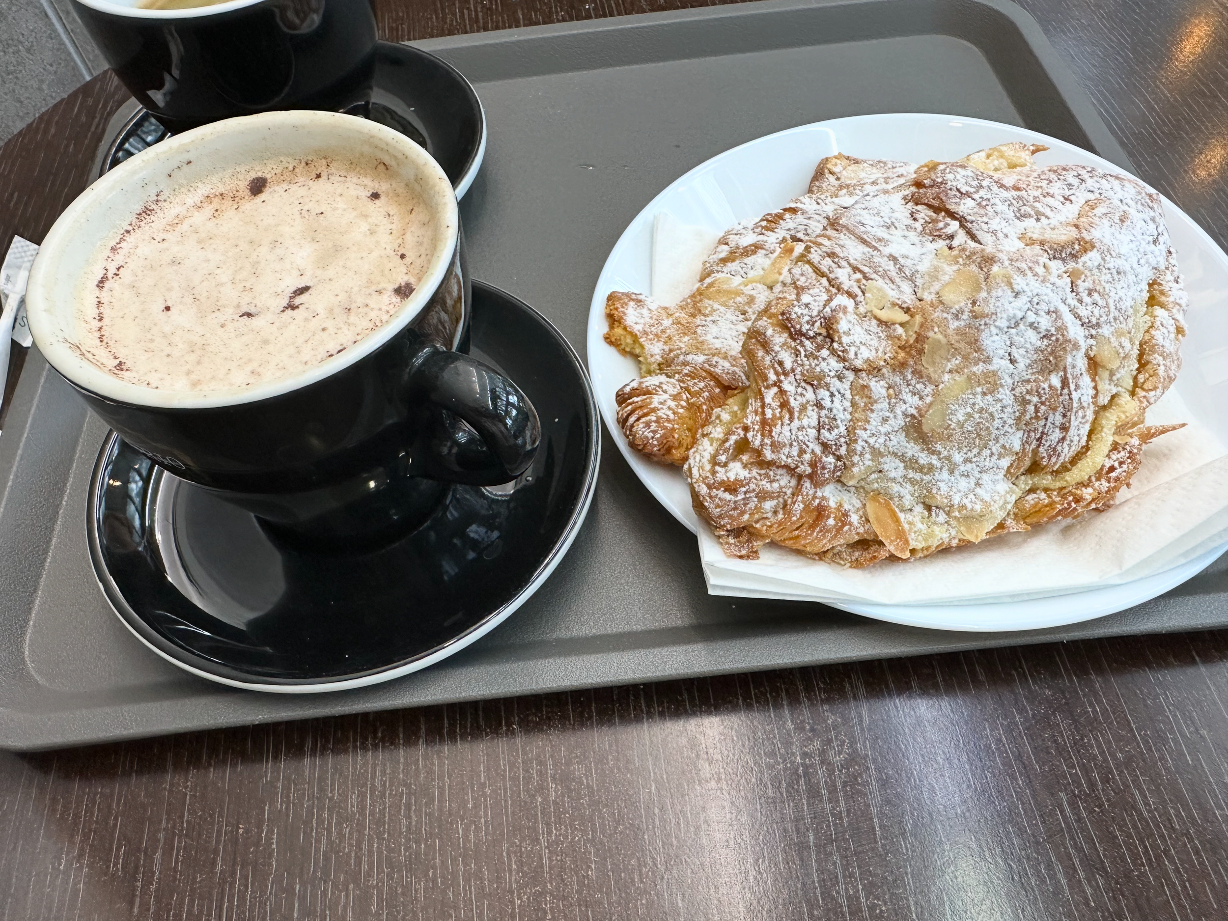 A cappuccino and a powdered-sugar almond croissant on a café tray.