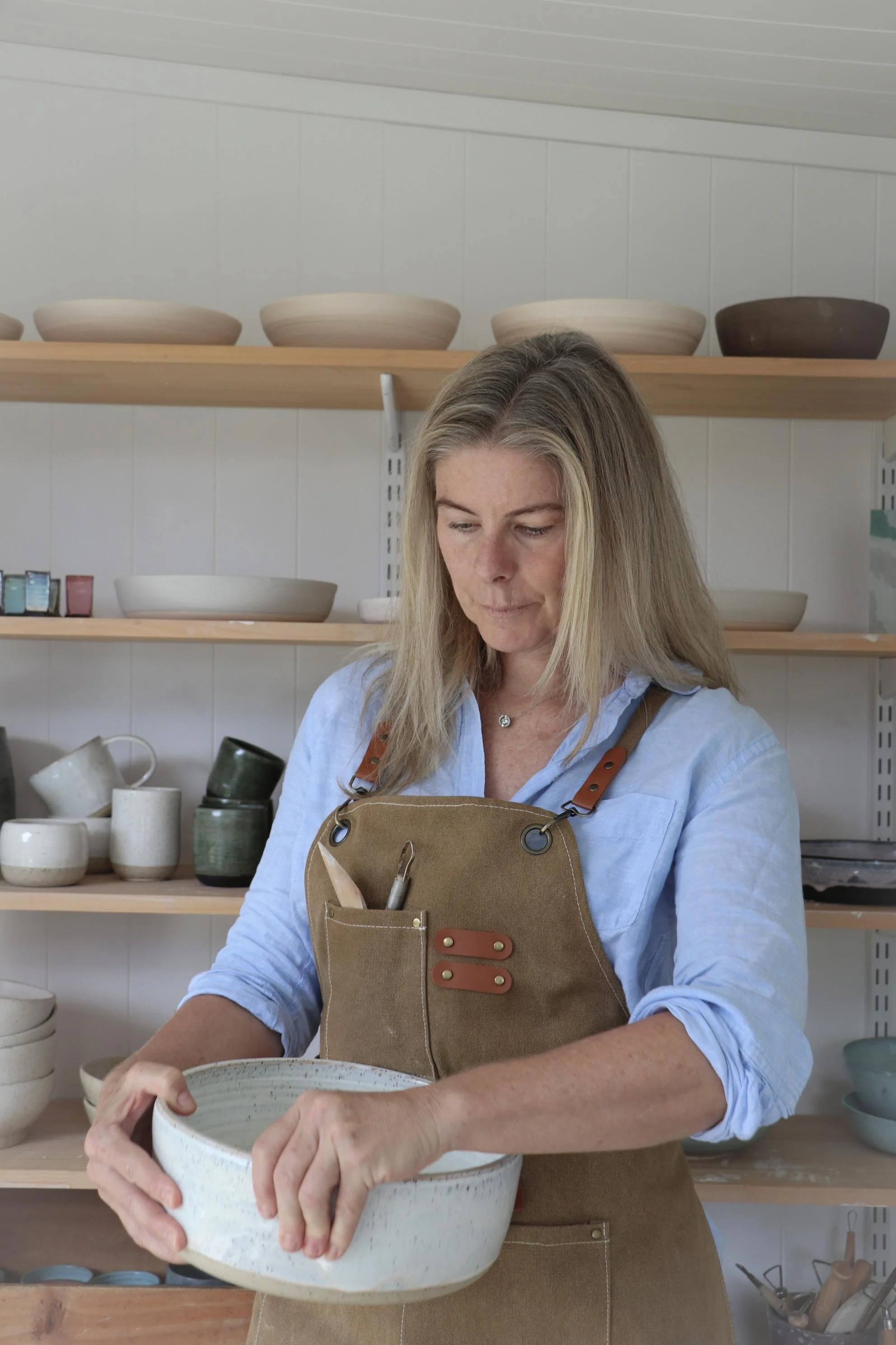 Gail Pettit, ceramic artist with blonde hair wearing a brown apron and light blue shirt, working in a ceramics studio, holding a large, speckled ceramic bowl.