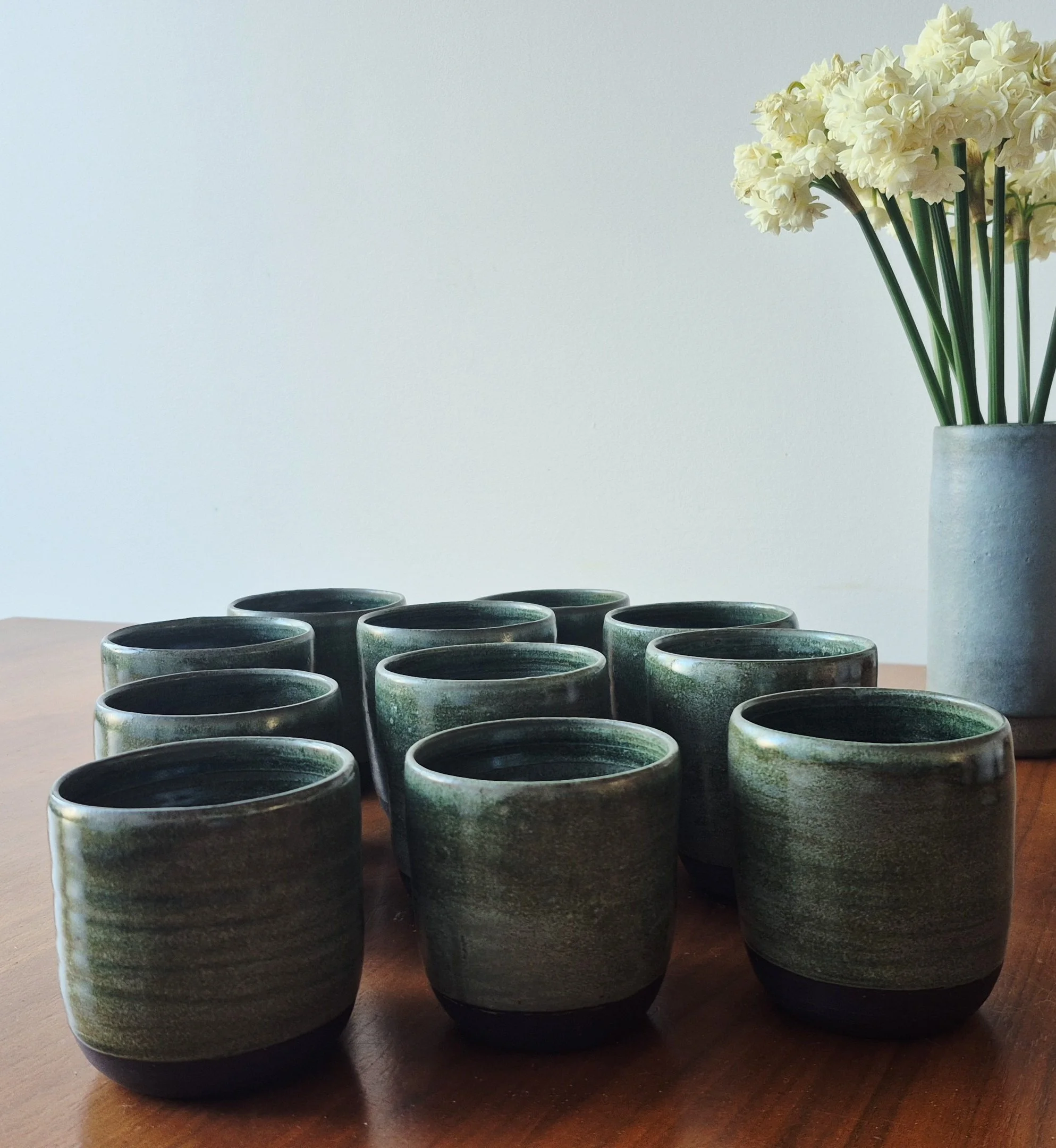 A collection of black and green ceramic cups arranged on a wooden surface with a white vase of white flowers in the background.