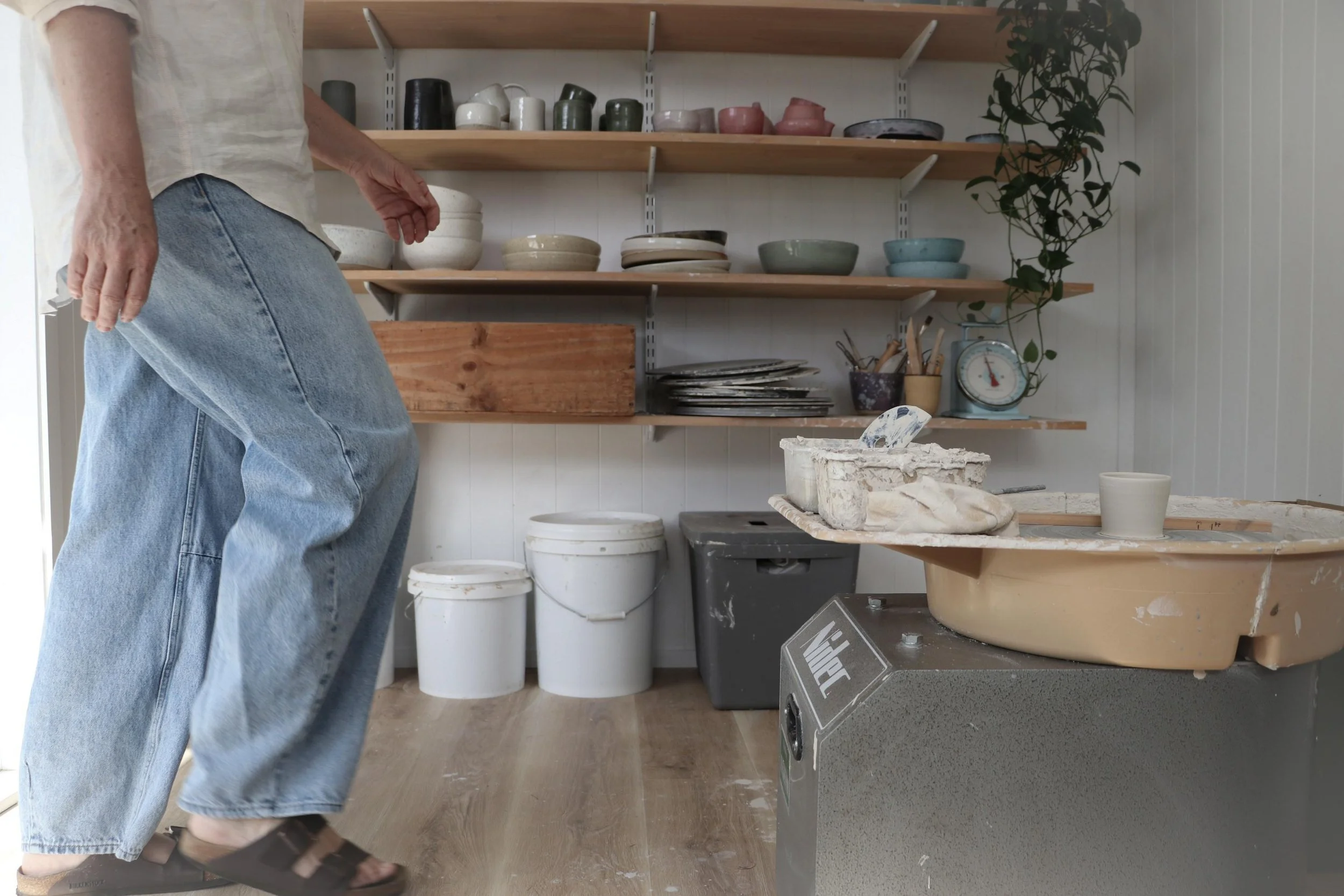 Pettit Ceramics,  wearing jeans and sandals working in a ceramics studio with shelves of ceramic bowls and cups, pottery tools, and a pottery wheel.