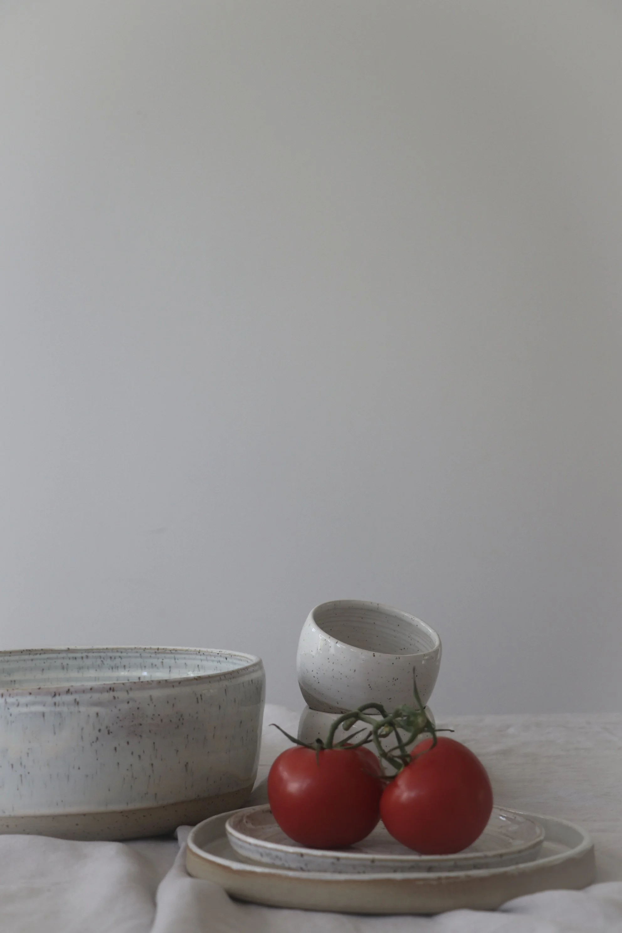Three white handmade ceramic bowls and a white ceramic plate with three tomatoes on it, set against a white background made by New Zealand ceramic artist and founder of Pettit Ceramics, Gail Pettit.