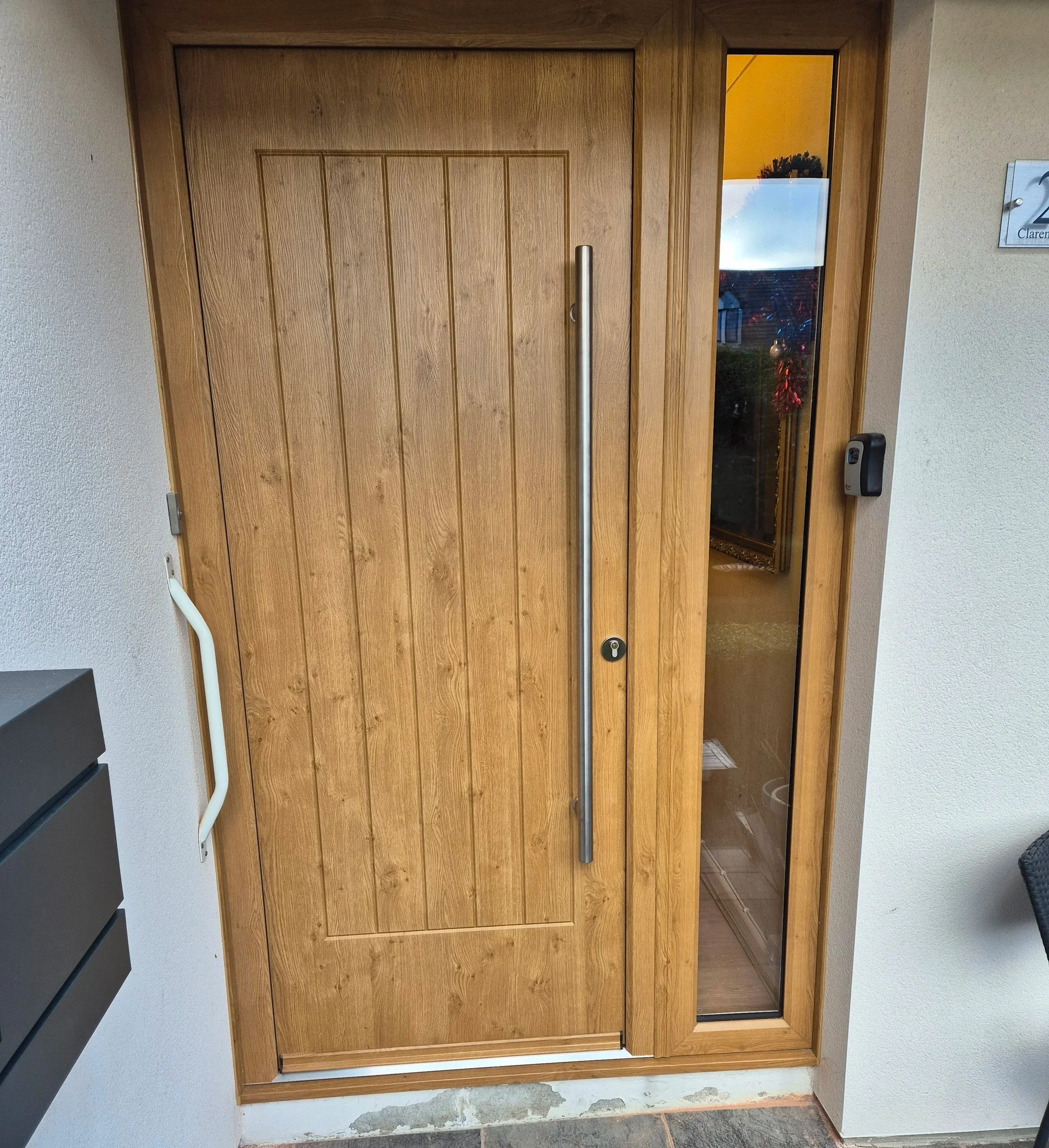 Oak composite front door featuring a realistic wood-grain cottage panel design and a matching full-height glazed side panel. This contemporary installation is finished with a sleek vertical bar handle, delivering a premium look with high thermal effi