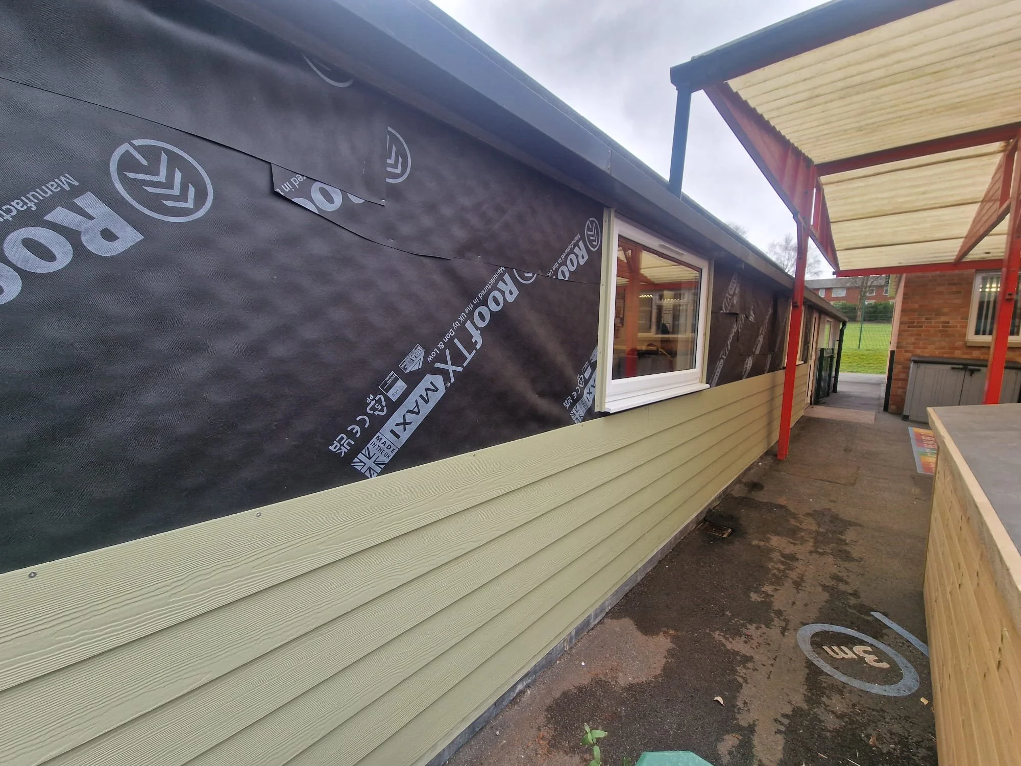 School cladding installation, with multi-layer thermal and weatherproofing system. High-density insulation and protective membrane layers installed beneath the durable sage green composite cladding, engineered to drastically improve the school buildi