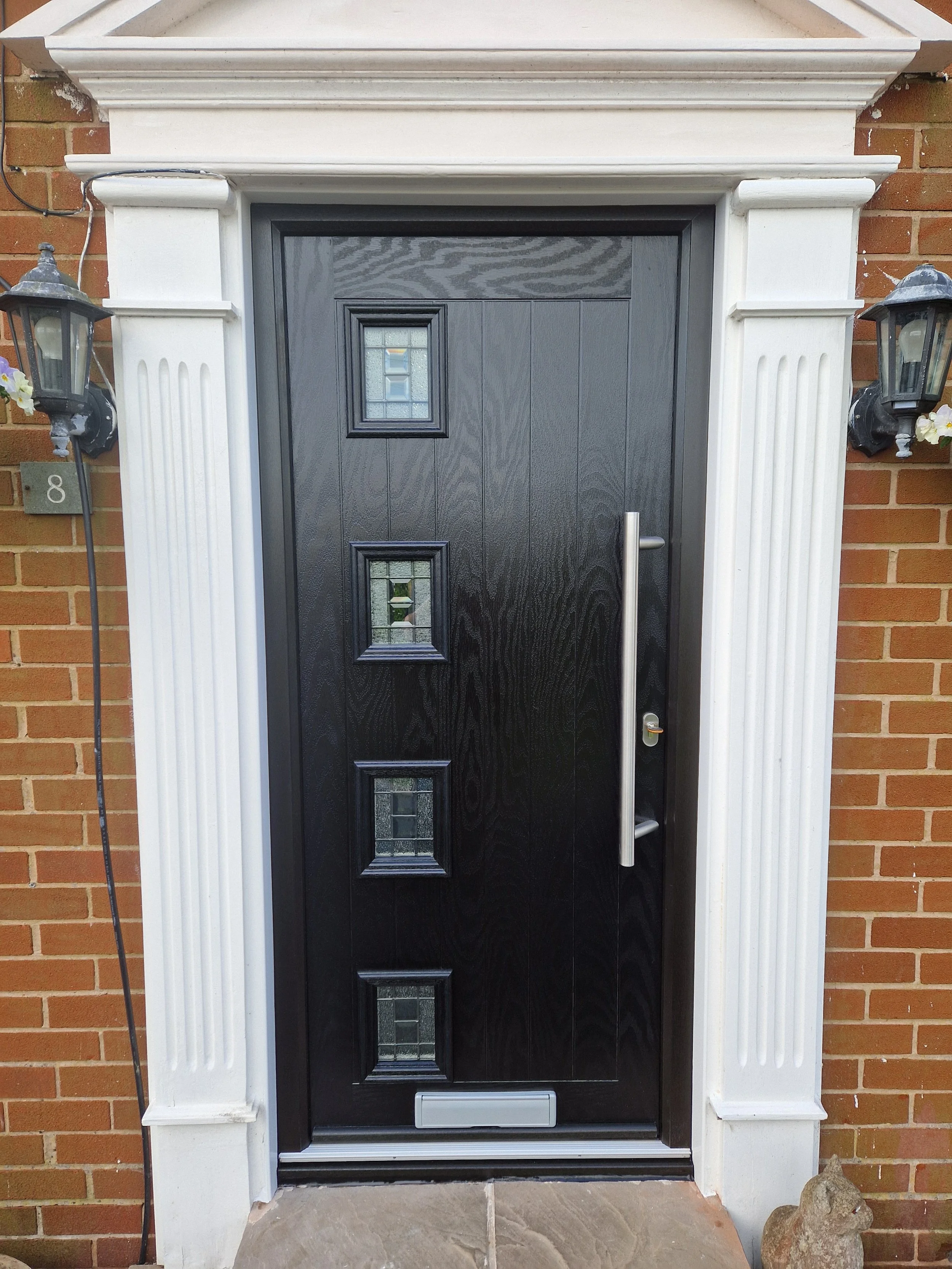 Premium black composite front door featuring a sleek vertical bar handle and four decorative glass panels. The sophisticated design and crisp white trim deliver a prestigious, high-end appearance.
