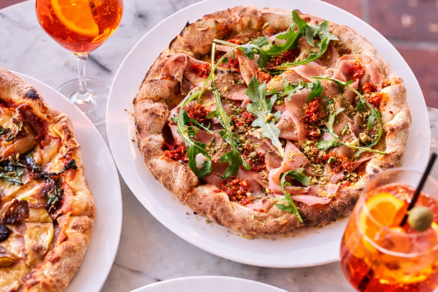 A pizza topped with arugula, prosciutto, crushed pistachios, and red pepper flakes served on a white plate, with a glass of orange-colored drink with a slice of orange and olive garnish, on a marble surface.