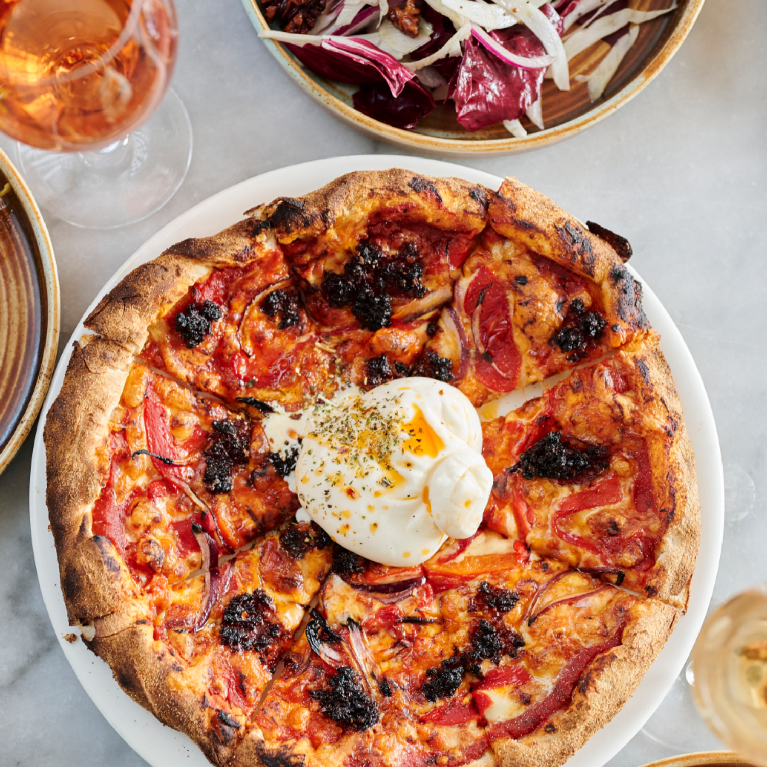 Pizza with burrata on top, served on a white plate, with a bowl of salad nearby and glasses of rosé in the background.