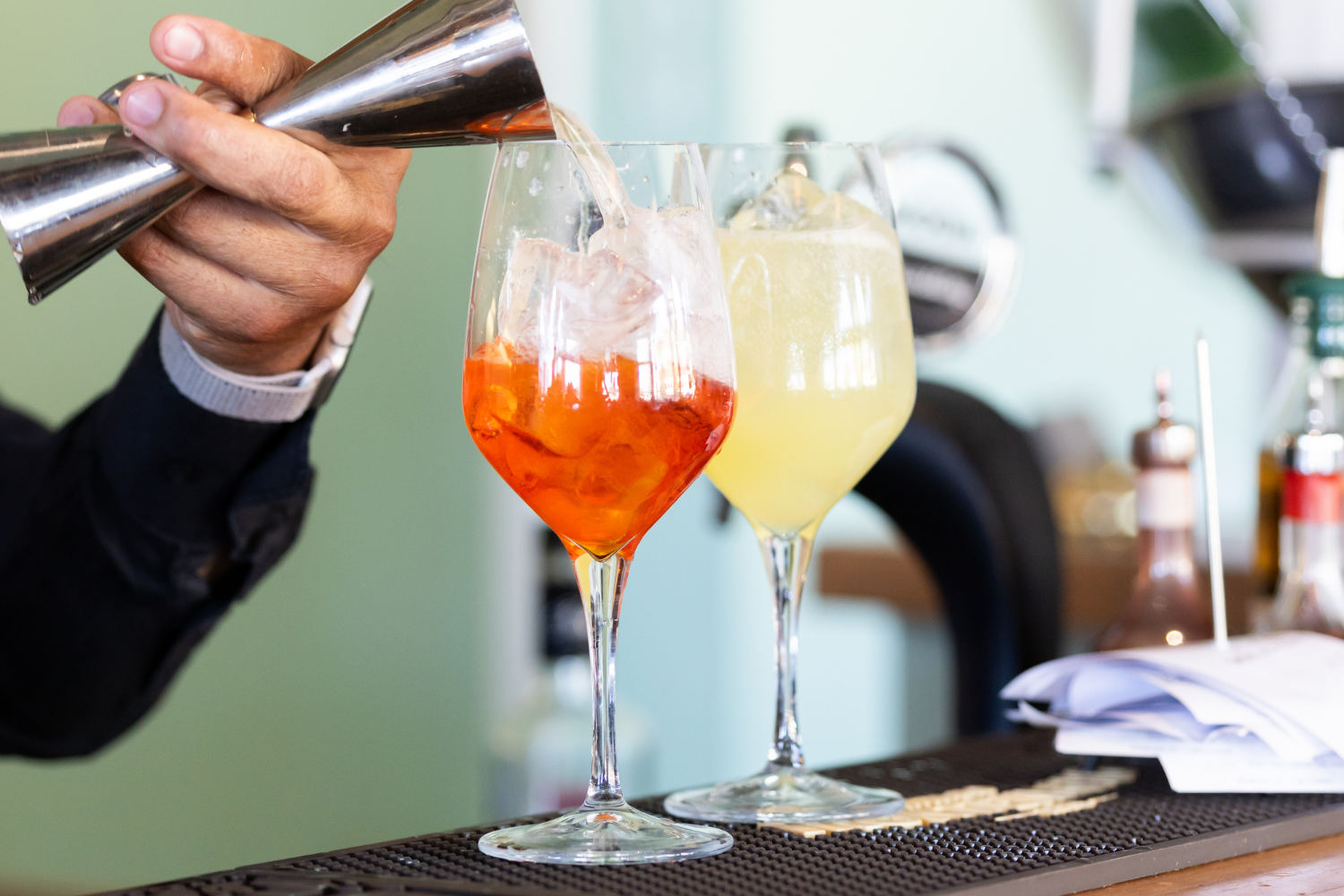 A bartender pours a cocktail into a glass with ice and orange liquid, with another yellow cocktail next to it, on a bar counter.
