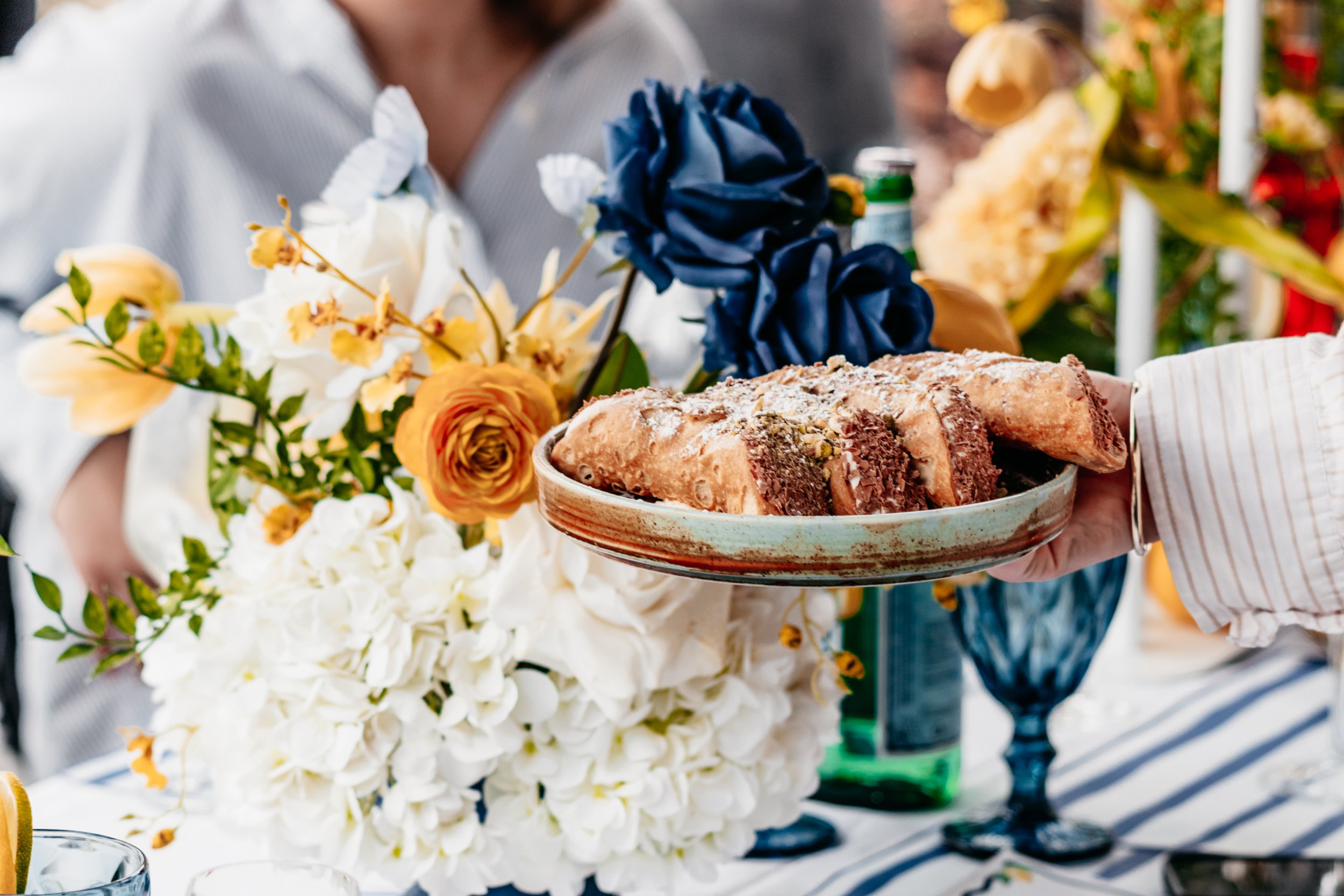Person holding a plate with sliced dessert next to a floral arrangement on a table at a celebration.