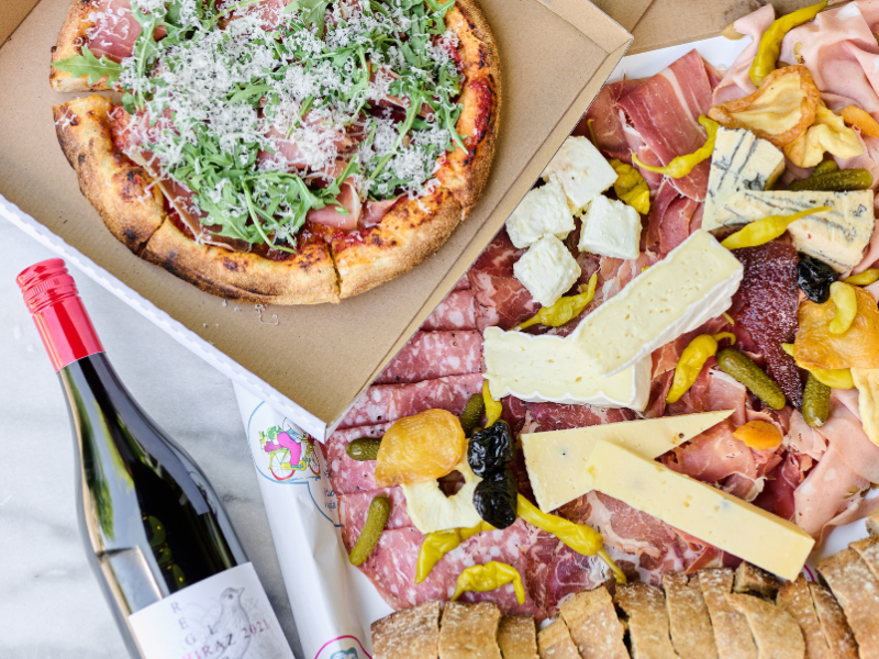 A pizza with arugula, prosciutto, grated cheese, and tomato sauce; a charcuterie board with various cheeses, cured meats, pickles, olives, and bread; and a bottle of red wine.
