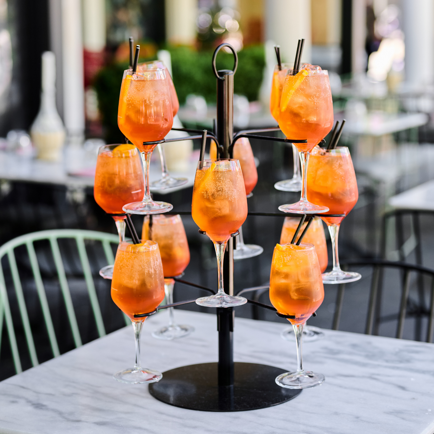 A tiered black metal stand holding twelve glasses of orange-colored cocktails, garnished with lemon slices and black straws, on a white marble table outdoors.