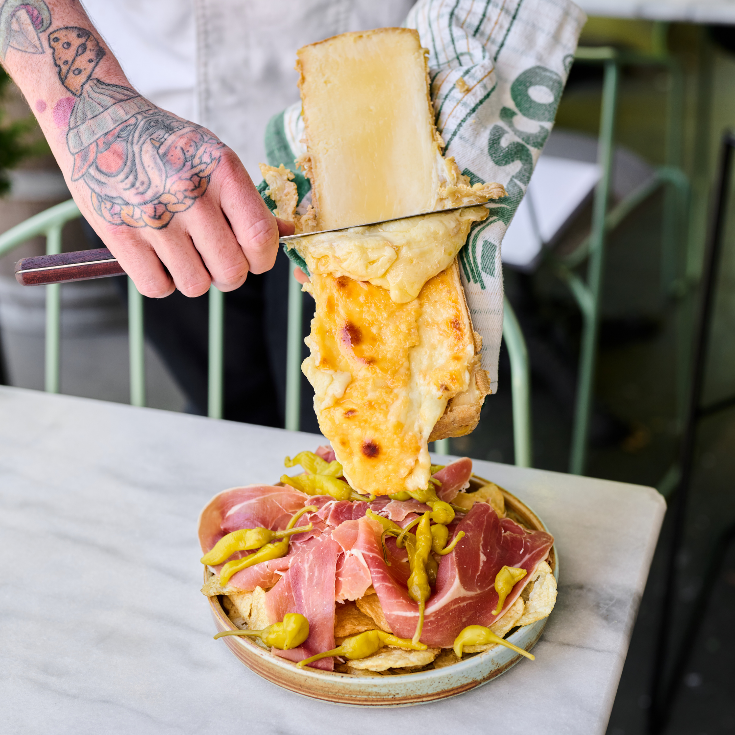 A person with tattooed arm slicing raclette onto a plate of cured ham and green peppers, with a marble table surface in the foreground.