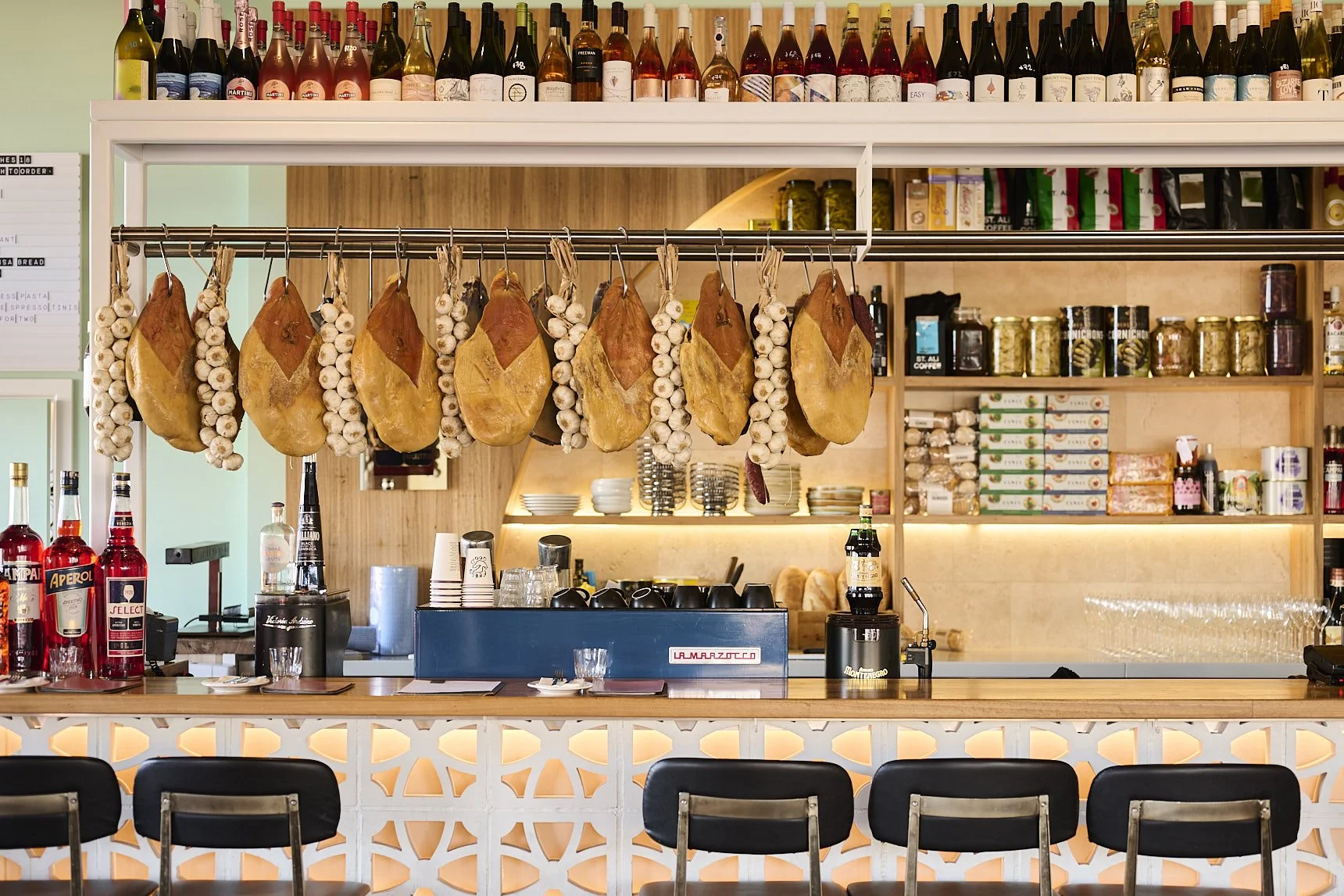 A bar with hanging cured ham and garlic bulbs above the counter, shelves stocked with bottles of alcohol, jars, and boxes, and bar equipment, with stools in front.