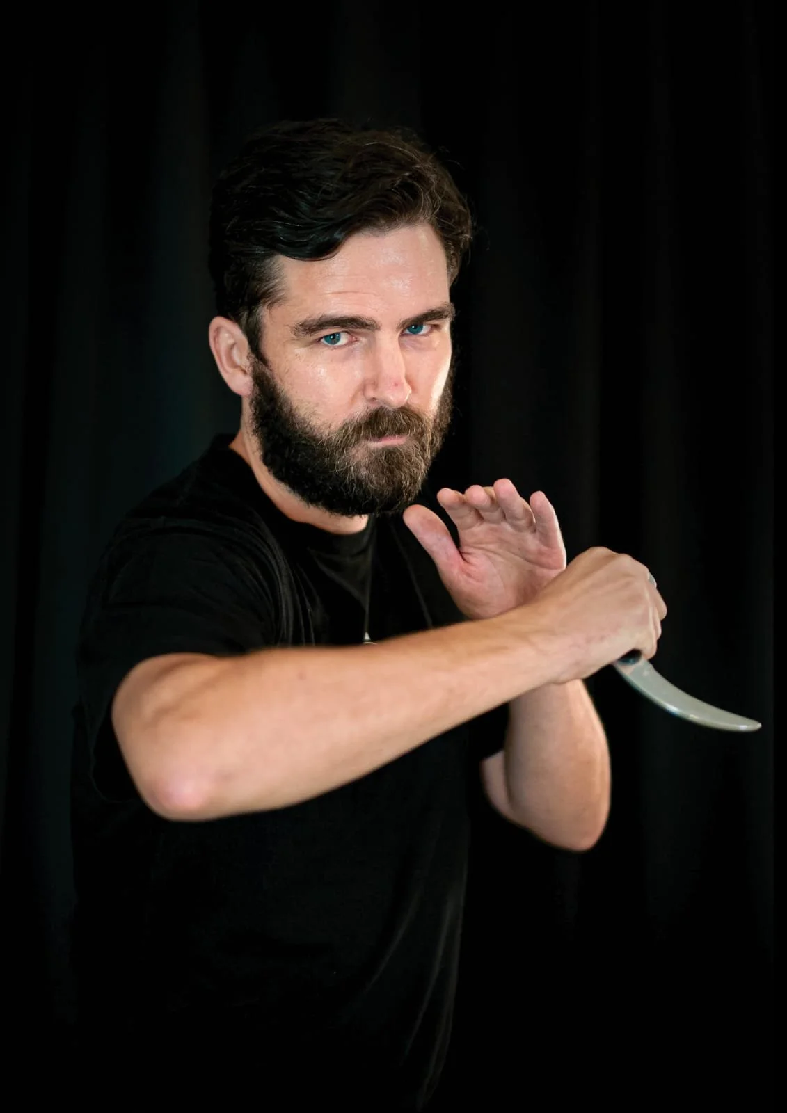 A man with dark hair and a beard holding a curved knife in a threatening pose against a black background.