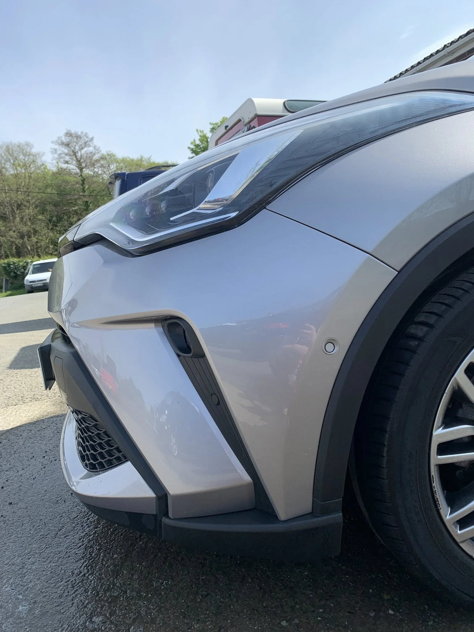 Close-up of the front side of a silver modern car, showing the headlight, bumper, and front wheel, with a background of trees, a blue sky, and other parked vehicles.