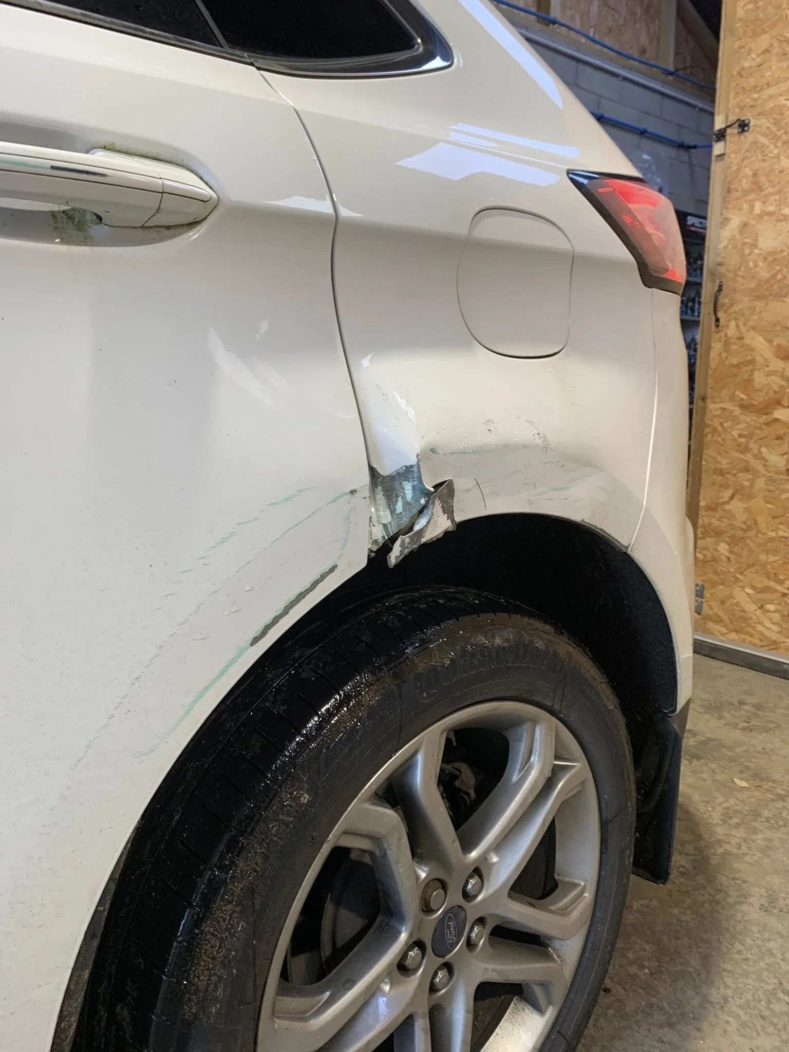 White car with visible damage to the rear side panel near the wheel, showing dents and scratches.